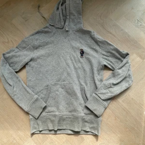 Grå hoodie Polo Bear från Ralph Lauren - Säljer en grå hoodie från Ralph Lauren med Polo Bear-broderi på bröstet. Tröjan har huva med dragsko, känguruficka och långa ärmar. Mjuk bomullskänsla och klassisk passform, perfekt för chill dagar. Tveka inte på att höra av dig!!!