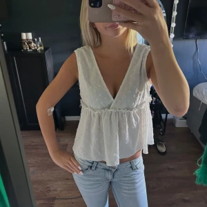 Vit blommig ärmlös blus med v-ringning - Supersöt vit blus med broderade små blommor, djup v-ringning och volang nedtill. Ärmlös modell med luftig passform, perfekt för varma dagar. Tunt och lätt tyg som ger en romantisk vibe.