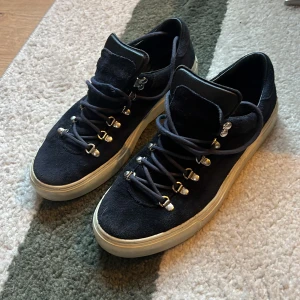 Mörkblå mockasneakers från Diemme - Snygga mörkblå sneakers från Diemme i mocka med robusta metalldetaljer och snörning. Skorna har vit platt sula och en unik design med grova öglor för snörena. Insidan är fodrad med svart skinn för extra komfort. Perfekta för dig som gillar stilrena men ändå sportiga skor. Fint men använt skick. Endast skor ingen låda