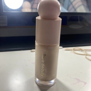 Rare Beauty flytande highlighter - Superfin flytande highlighter från Rare Beauty i en ljus champagnefärg. Kommer i en snygg glasflaska med ljusrosa lock och rund topp, perfekt för att ge glow till kindbenen eller andra partier i ansiktet.