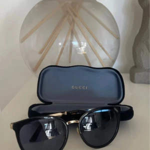 Svarta solglasögon från Gucci - Äkta! Snygga svarta solglasögon från Gucci med rundad form och gulddetaljer vid skalmarna. Modellen är GG0077SK och de har mörka glas. Kommer med originalfodral. Perfekta för att lyfta din stil i sommar. OBS! Äkta gucci glasögon inköpt från synsam  OBS! Den har styrkor på -1.00. Ni som inte har syn problem kan uppleva att man bli snurrig av den då den har svag styrka på båda. Knappt använd. Självklart kan man byta glasrutorna för att få det att passa.