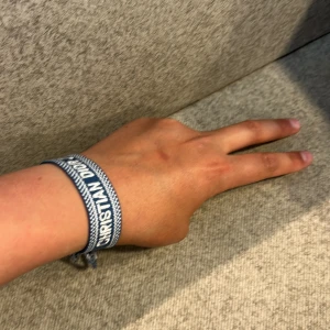 Blått Christian Dior J'ADIOR armband - Snyggt blått tygarmband från Christian Dior med texten J'ADIOR i vitt. Armbandet har flätade band och tofsar i ändarna för en cool och avslappnad look. Perfekt accessoar för att lyfta din outfit med en touch av lyx.