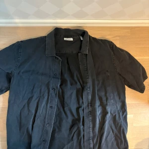 Svart overshirt från Weekday - Svart overshirt från Weekday i bomull med kort ärm och klassisk krage. Skjortan har knappar framtill och en bröstficka. Perfekt för en avslappnad och stilren look.