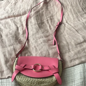 Rosa axelväska från H&M med flätad botten - Snygg axelväska från H&M med rosa lock och axelrem i syntet, samt flätad beige botten. Väskan har guldfärgad ringdetalj framtill och innerfack med dragkedja. 20cm bred 15cm höjd. 