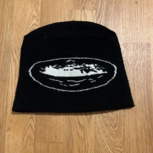 Svart mössa med vit grafik - Svart mössa i klassisk modell med vit grafisk design framtill som föreställer ett fartyg i en oval ram. Enkel och stilren, perfekt för kalla dagar. Materialet är stickat och ger en mjuk känsla mot huvudet.