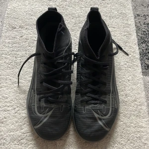Nike Mercurial svarta fotbollsskor - Svarta Nike Mercurial fotbollsskor med högt skaft och snörning. Snygg design med silvriga detaljer och Nike-logga på sidan. Ovandel i syntetmaterial och greppvänlig sula för bra kontroll på planen. Perfekta för dig som vill sticka ut på futsal eller konstgräs.