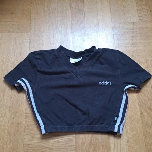 Svart kort topp från Adidas - Svart kort topp från Adidas med v-ringning och korta ärmar. Snygga vita ränder längs sidorna och broderad Adidas-logga framtill. Storlek D36 / 38. 