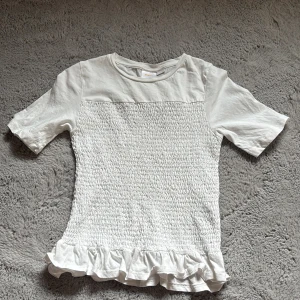 Vit blus med volang från Gina Tricot - Supersöt vit blus från Gina Tricot med smockad framsida och volangkant nertill. Toppen har korta ärmar och rund halsringning, perfekt för en fräsch och trendig look. 