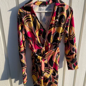 Färgstark omlottklänning från Boohoo - Säljer en kort omlottklänning från Boohoo med färgstarkt mönster i svart, gul, rosa och röd. Klänningen har långa ärmar, v-ringning och knyt i midjan. Perfekt för dig som gillar att sticka ut med coola retrovibbar och unika detaljer.