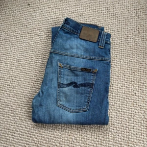 Blå jeans från Nudie Jeans - Säljer ett par klassiska blå jeans från Nudie Jeans med snyggt broderad bakficka och läderpatch i midjan. Jeansen har en rak passform och är tillverkade i mjukt denimtyg. Perfekta för dig som gillar stilrena och tidlösa jeans. En liten defekt i en av fickorna men Nudie erbjuder gratis reparation på sina byxor.