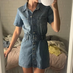 Jeansklänning med flätat bälte - Snygg jeansklänning i blå denim med kort ärm, knappar hela vägen framtill och två bröstfickor. Midjan markeras av ett flätat bälte i samma material. Klassisk krage och avslappnad passform, perfekt för en cool och avslappnad look.