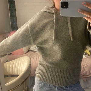 Grå stickad hoodie med huva - Mysig grå stickad hoodie med huva och snörning. Tröjan har breda ribbade muddar vid ärmslut och midja, samt en avslappnad passform. Perfekt för kyliga dagar när du vill vara både bekväm och snygg.
