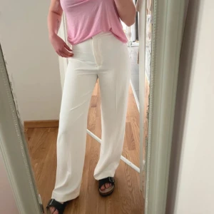 Vita vida kostymbyxor med hög midja - Säljer ett par stilrena vita kostymbyxor med hög midja och raka, vida ben. Byxorna har en klassisk look och är tillverkade i ett lätt tyg som ger en snygg siluett. Perfekta att matcha med sneakers eller sandaler för en fräsch vibe.