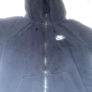 Svart Nike hoodie med dragkedja - Svart hoodie från Nike med vit broderad logga på bröstet. (Hål bakom axeln typ visar på bilden men annars i bra skick den e knappt använd skulle säga att den passar S också  