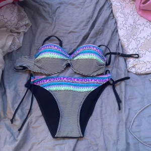 Randig bikini med färgglada detaljer - Snygg bikini från FUBA. VI med svartvita ränder och färgglada textdetaljer i blått, rosa och grönt. Överdelen har bygel och justerbara axelband, underdelen har knyt i sidorna. Perfekt för stranden eller poolen!