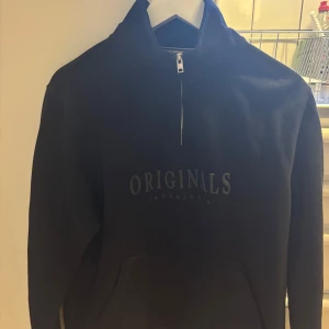 Svart half zip hoodie Jack & Jones - Svart hoodie från Jack & Jones med trycket 'ORIGINALS' på bröstet. Tröjan har half zip-dragkedja i halsen, känguruficka och hög krage. Väldigt lite använd 
