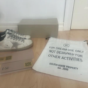 Golden Goose Superstar sneakers vit/grå - Golden Goose Superstar sneakers i vitt skinn med grå stjärndetalj på sidan, grå snörning och svart häl. Klassisk låg modell med rund tå och platt sula. Snyggt slitna detaljer för en cool och trändig-look. Kommer med originalpåse och låda. Säljes pga att de var för små. Jag har använt skorna en två gånger men annars är dem helt nya. Det är bara att höra av sig vid funderingar.