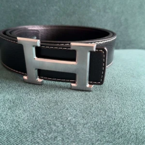 Svart bälte, skärp med H-spänne - Snyggt svart skärp i silverfärgad metall. Bältet är gjort i äkta skinn och har vita kontrastsömmar längs kanterna. Perfekt accessoar för att lyfta din outfit med en lyxig touch.