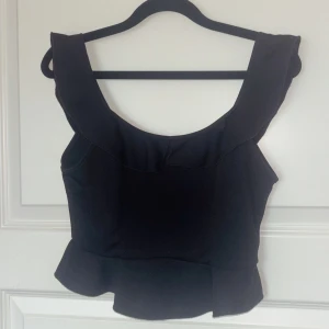 Svart croppad topp från Bik Bok - Snygg svart croppad topp från Bik Bok med breda axelband och volangdetalj nedtill. Toppen har en enkel och stilren design som passar perfekt till jeans eller kjol. Materialet känns mjukt och följsamt mot kroppen. Nästan aldrig använd så inga skavanker finns. OBS tryck ej på köp direkt då jag har lagt ut på flera ställen! Skriv till mig innan💕