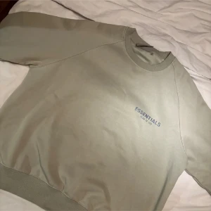 Beige Essentials Fear of God sweatshirt - Snygg beige/grön sweatshirt från Essentials Fear of God med diskret logga på bröstet och ärmen. Tröjan har rund hals, ribbade muddar och är tillverkad i mjuk bomullsmix. Perfekt för en clean och avslappnad streetwear-look. Storlek s men upplevs mer som medium, aldrig använd