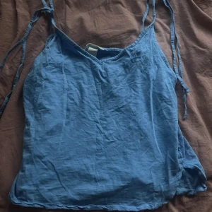 Blå linnetopp med knytband - Säljer en enkel blå linnetopp i storlek M med smala axelband som knyts upptill. Toppen har en avslappnad passform och är tillverkad i ett lätt material, perfekt för varma dagar. Snygg att matcha med jeans eller shorts för en chill look.