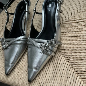 Silvriga spetsiga pumps från Zara - Silvriga pumps från Zara med spetsig tå och flera smala remmar med små spännen över foten. Skorna har öppen häl och justerbara remmar runt vristen. Snygg metallic-finish som verkligen sticker ut och ger en futuristisk vibe.