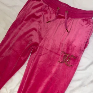 Rosa velourbyxor Juicy Couture - Säljer ett par ikoniska rosa mjukisbyxor från Juicy Couture i mjuk velour. Byxorna har snörning i midjan och glittrande JC-logo i strass på benet. Perfekta för en chill och trendig look.