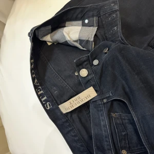 Svarta jeans från Burberry - Snygga svarta jeans från Burberry med klassisk femficksdesign och diskret metallogo vid fickan. Jeansen har mörk tvätt och raka ben, samt rutigt innerfoder i fickorna som ger en exklusiv touch. Perfekta för dig som gillar stilrena och tidlösa plagg.