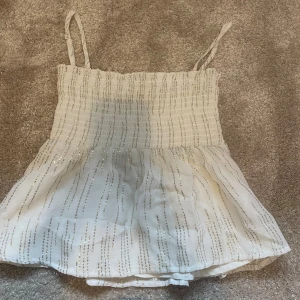Vit glittrig linnetopp från Gina Tricot - Supersöt vit linnetopp från Gina Tricot med smala axelband och glittriga silverränder. Axelbanden går att ta bort så man kan ha den som en kjol eller en topp. Toppen har smockad överdel och ett lätt utställt fall. Perfekt för dig som vill ha något extra till sommaren!
