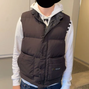 Svart dunväst från Canada Goose - Säljer en svart dunväst från Canada Goose med hög krage och stora fickor framtill. Västen har både dragkedja och knappar, samt en diskret logga på ena fickan. (Modellen säljs inte längre)