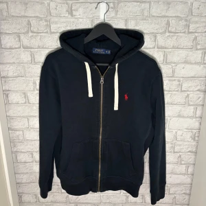 Ralph Lauren zip - Svart Ralph Lauren hoodie med vit snörning i huvan och dragkedja framtill. Klassisk röd logga broderad på bröstet och två stora fickor. Tröjan är i mycket fint skick utan tecken på användning! Hör av dig vid intresse 🙌