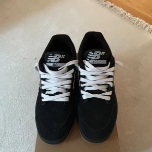 New Balance 480 svarta sneakers - Svarta New Balance 480 sneakers med vit logga på sidan och vita snören. Ovandel i mocka och syntet, klassisk låg modell med gummisula i brun ton. Perfekta för dig som gillar stilrena och sportiga skor med retrovibbar. Kommer med svarta skosnören också!