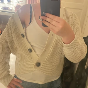 Beige stickad kofta med stora knappar - Säljer en beige stickad kofta med stora svarta knappar. Koftan har långärmade ärmar med breda muddar. Perfekt att slänga över ett linne eller t-shirt för en chill och snygg look.