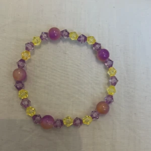 Färgglatt pärlarmband i lila och gult - Ett stretchigt armband med glittriga, facetterade pärlor i lila och gult samt större runda pärlor i lila och orange. Perfekt accessoar för att liva upp din outfit med färg och lekfullhet.