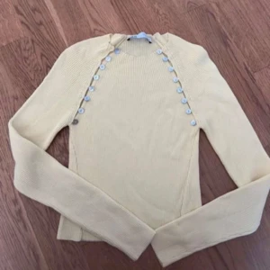 Gul ribbad tröja med knappar från Zara - Supersöt ljusgul ribbad tröja från Zara med långa ärmar och snygga knappar längs axlarna. Tröjan är figurnära och har en mjuk, stickad känsla som passar perfekt till jeans eller kjol. Enkel men ändå unik detalj med knapparna framtill.