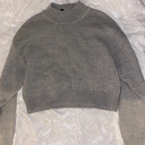 Grå stickad croppad tröja - Mysig grå stickad tröja med croppad passform och hög krage. Perfekt för lager-på-lager och passar till både jeans och kjol. Långärmad och ribbad vid ärmslut och nederkant för extra stilpoäng.