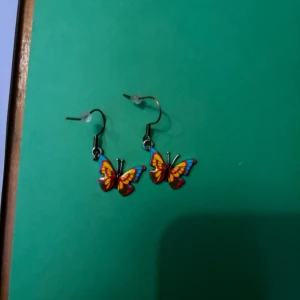 Färgglada fjärilsörhängen - Säljer ett par superfina örhängen med hängande fjärilar i rött, gult och blått. Perfekta för dig som vill ha en färgklick och lekfull stil. Fjärilarna har tydliga detaljer och örhängena har klassisk krok. Passar dig som gillar unika accessoarer.