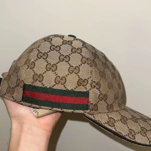 Gucci keps med GG-mönster och röd/grön detalj - Priset är fast och om ni har flera frågor eller om ni är intresserade hör av er.                                  Snygg keps från Gucci i beige och brunt med klassiskt GG-monogram över hela kepsen. Har en ikonisk röd och grön rand på sidan och justerbar rem i brunt skinn baktill. Tillverkad i Italien och perfekt för dig som vill sticka ut med lyxig streetstyle.