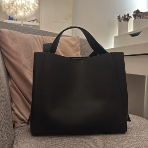 Svart stilren axelväska i skinn - Minimalistisk svart axelväska i slätt skinn med breda handtag och stilren design. Väskan har en rymlig insida och en innerficka med dragkedja för extra säkerhet. Perfekt för dig som gillar en clean och modern look. Från ginatricot, helt slutsåld. 