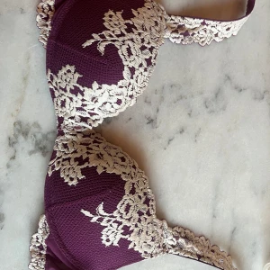 Lila spets-bh med blommönster - Säljer en lila bh med vacker vit spets och blommigt mönster. Bh:n har vadderade kupor och breda axelband dekorerade med spets. Perfekt för dig som vill ha något både snyggt och bekvämt.