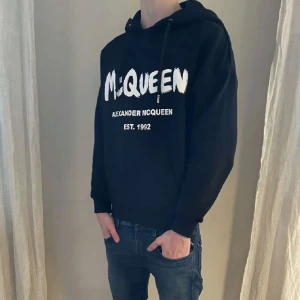 Svart hoodie från Alexander McQueen - Säljer en stilren svart hoodie från Alexander McQueen med vit logotyp och text 'EST. 1992' på framsidan. Hoodien har en klassisk design med dragsko i huvan och ribbade muddar vid ärmslut och nederkant. Perfekt för en avslappnad look. Hör av dig vid frågor eller funderingar.