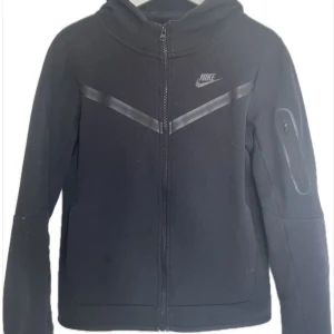 Svart zip-hoodie från Nike - Svart hoodiejacka från Nike med dragkedja framtill och huva. Jackan har en diskret logga på bröstet, svarta detaljer i form av ett V över bröstet och en ficka med dragkedja på ena ärmen. Perfekt för en sportig look.