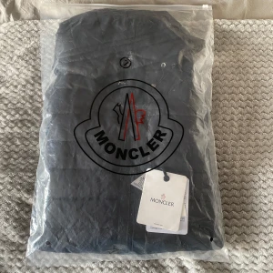 Svart pufferjacka från Moncler med huva - Säljer en svart pufferjacka från Moncler med stickade ärmar och vadderad kropp. Jackan har huva, dragkedja framtill och två fickor med dragkedja. Klassisk Moncler-logga på ärmen och snygga detaljer. Perfekt för kyliga dagar. Storlek M men passar även S , 