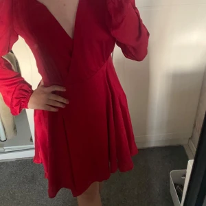 Röd omlottklänning med v-ringning - Säljer en röd omlottklänning med v-ringning och långa ärmar. Klänningen har en kort längd och är tillverkad i ett lätt, flowy material som ger en snygg siluett. Perfekt för dig som vill sticka ut med färg!