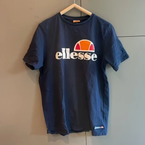 Blå t-shirt från Ellesse - Säljer en snygg blå t-shirt från Ellesse med deras ikoniska logga i vitt, orange och rött på bröstet. T-shirten är kortärmad och har en klassisk rund halsringning. Perfekt för en avslappnad stil.