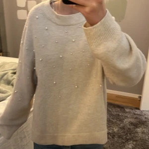 Kräm vit  stickad tröja med pärlor från H&M - Säljer en kräm-vit stickad tröja från H&M med dekorativa pärlor framtill. Tröjan har rund halsringning, långa ärmar och ribbade muddar vid ärmslut och nederkant. Perfekt för en mysig och stilren look!