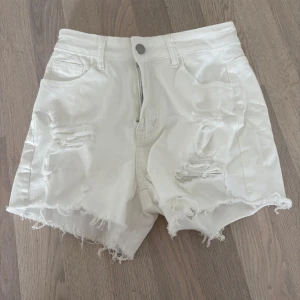 Vita jeansshorts med fransar - Säljer ett par vita jeansshorts med fransiga kanter och slitna detaljer. Shortsen har en dragkedja fram och är tillverkade av en bomullsblandning för extra komfort. Perfekta för en avslappnad sommarlook! Helt nya shorts endast testade men passade mig inte. Storlek M 