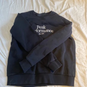 Svart sweatshirt från Peak Performance - Säljer en svart sweatshirt från Peak Performance med vit text på bröstet. Tröjan har långa ärmar och ribbade muddar vid ärmslut och nederkant. Perfekt för en avslappnad stil. Pris kan diskuteras
