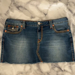 Jeanskjol från True Religion - Snygg jeanskjol från True Religion i en klassisk blå denimfärg. Kjolen har en kort längd med fransiga kanter och detaljerade bakfickor med knappar. Köpt för 1200kr och bara använd cirka 3 gånger.