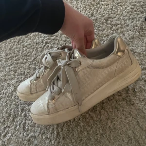 Beige sneakers med breda snören och gulddetaljer - Säljer ett par beige sneakers med breda snören och snygga gulddetaljer vid hälen. Skorna har rund tå och en tjock vit sula. Perfekta för dig som vill ha ett par stilrena och bekväma skor till vardags. Väldigt fint märke de brukar kosta kring 800-900kr nypris. Väl använda så lite små defekter tillkommer men inga man tänker, skriv för fler bilder eller mått. Svarar snabbt!